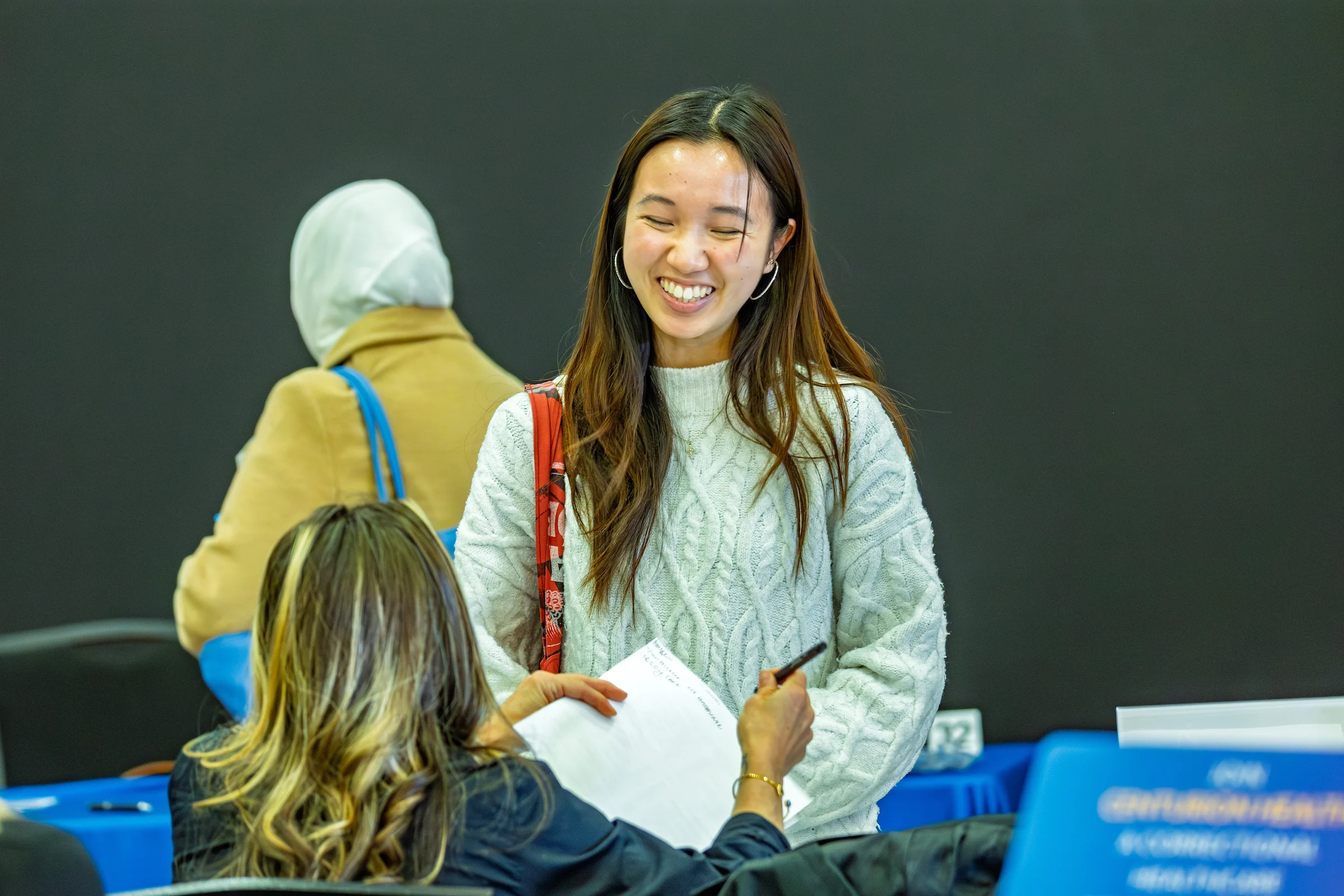 A woman smiles while holding papers, discussing career opportunities with another woman at a table during an indoor event.