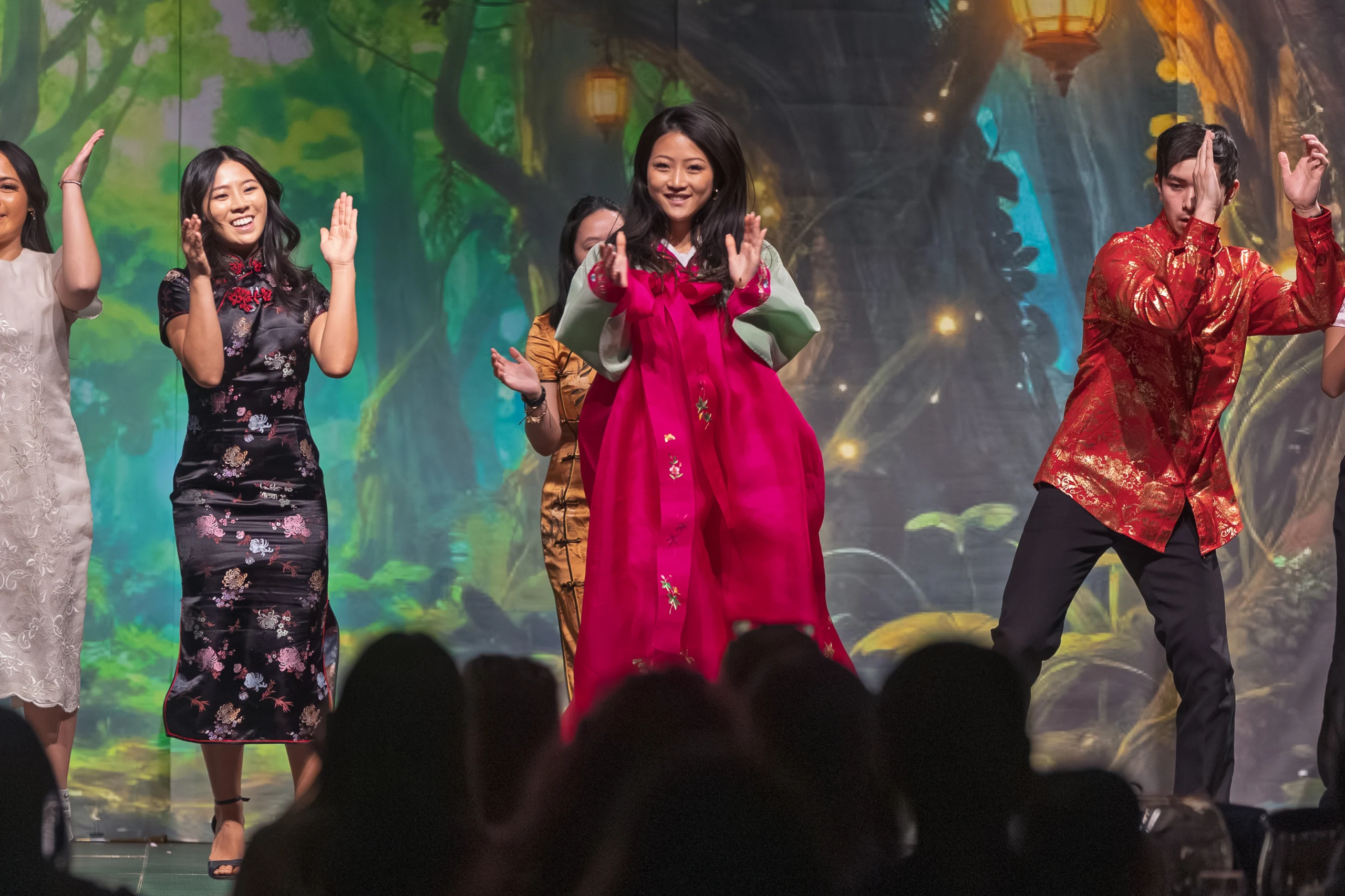 Students showcase their involvement as they dance joyfully on stage in colorful traditional and modern outfits, set against a vibrant forest-themed backdrop.