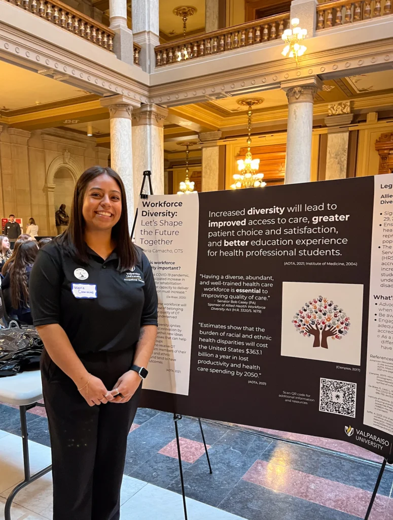 A woman stands smiling beside a display board about workforce diversity in a grand building interior, celebrating Valparaiso University graduates from the Occupational Therapy Doctorate program.