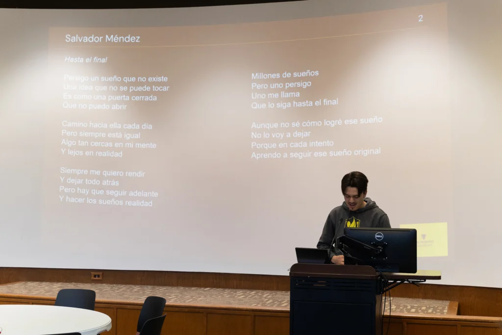 A student stands at a podium reading their Spanish poetry from Valpo's Letras Spanish Literary Magazine projected on a screen behind them.