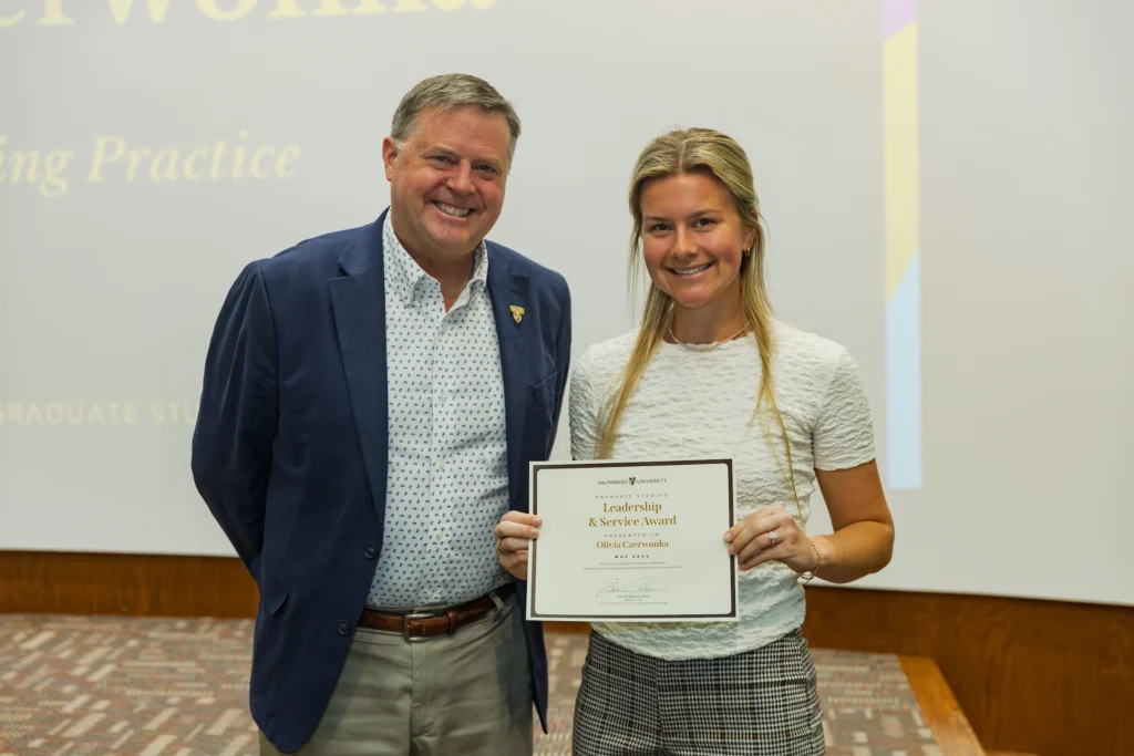 A young woman recognized for Graduate Student Excellence holds a certificate and stands smiling next to an older man in a suit at a Valpo Honors indoor event.