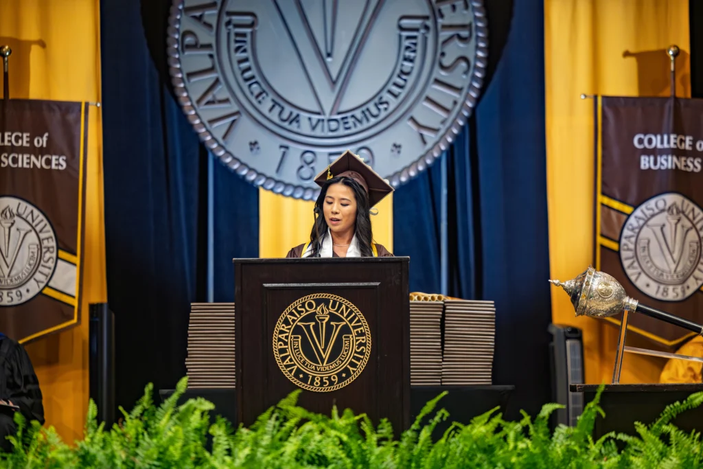 A graduate from the Valparaiso University graduating class of 2026 speaks at a podium during a commencement ceremony with banners and the university seal in the background.