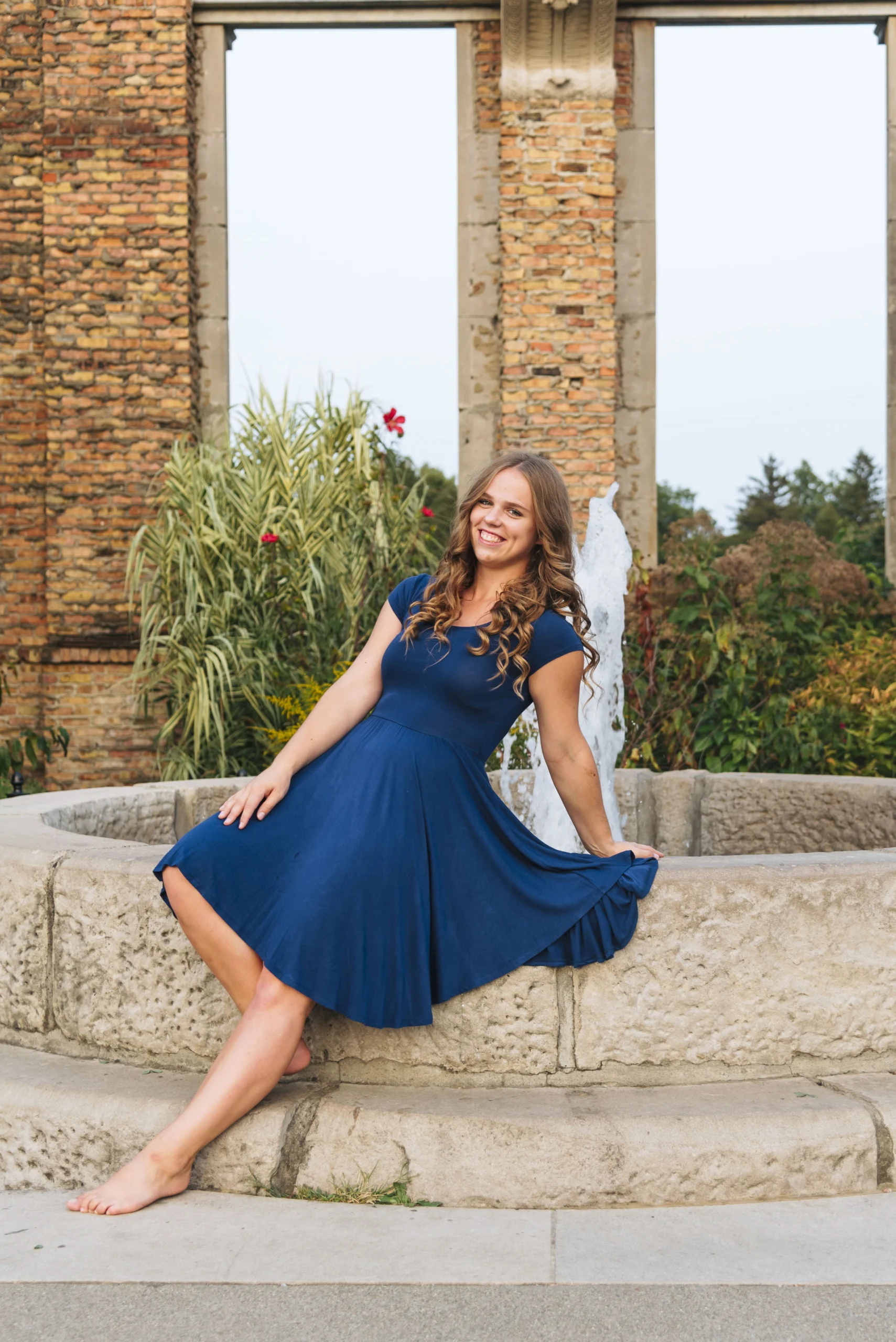 Smiling woman, Adele Beleckaite, in a blue dress sits barefoot on a stone fountain in a garden with brick pillars behind her, capturing the essence of Human Connection.