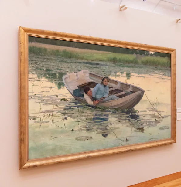 A framed painting of two people lying in a rowboat, floating on a lily-covered pond, hangs in the gallery as part of the Permanent Collection.