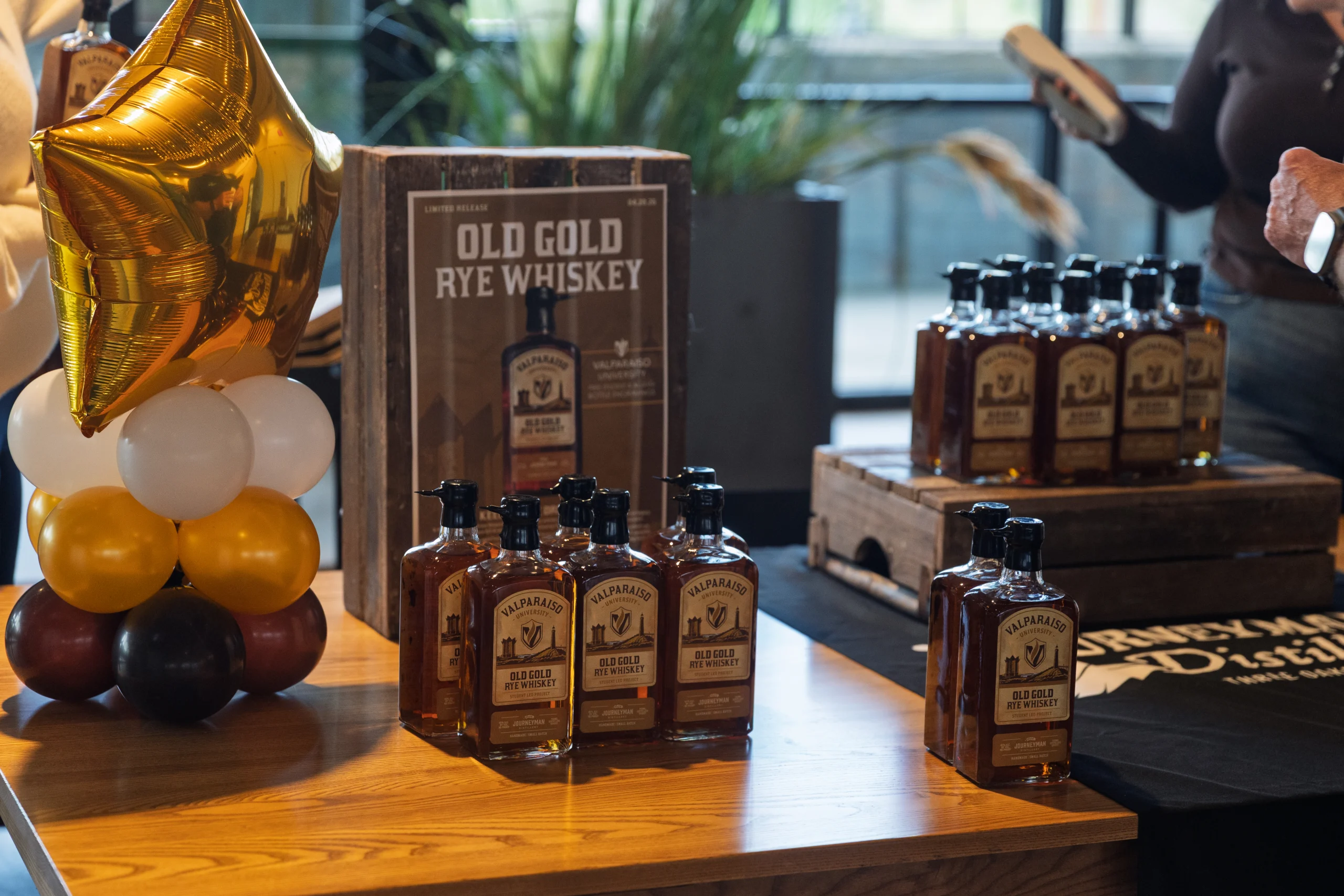 Bottles of Old Gold Rye Whiskey from Journeyman Distillery displayed on tables with a gold star balloon and festive decorations, celebrating Valparaiso University student entrepreneurs.