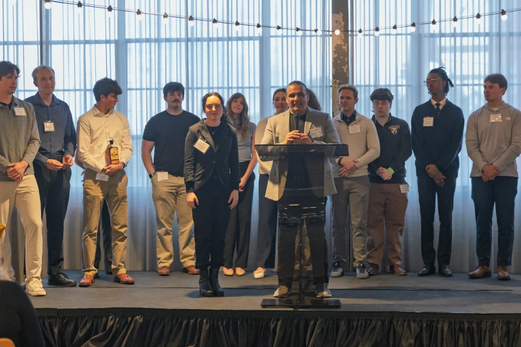 A group of student entrepreneurs from Valparaiso University stand on a stage at Journeyman Distillery, with one person speaking at a podium and string lights overhead.