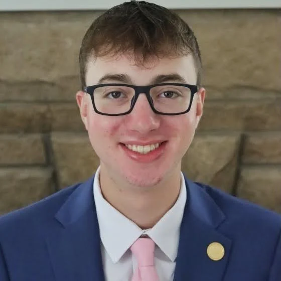 Young man in glasses, blue suit, white shirt, and pink tie, smiling in front of a stone wall—representing Valparaiso University at SORCE 2026 for Student Research.