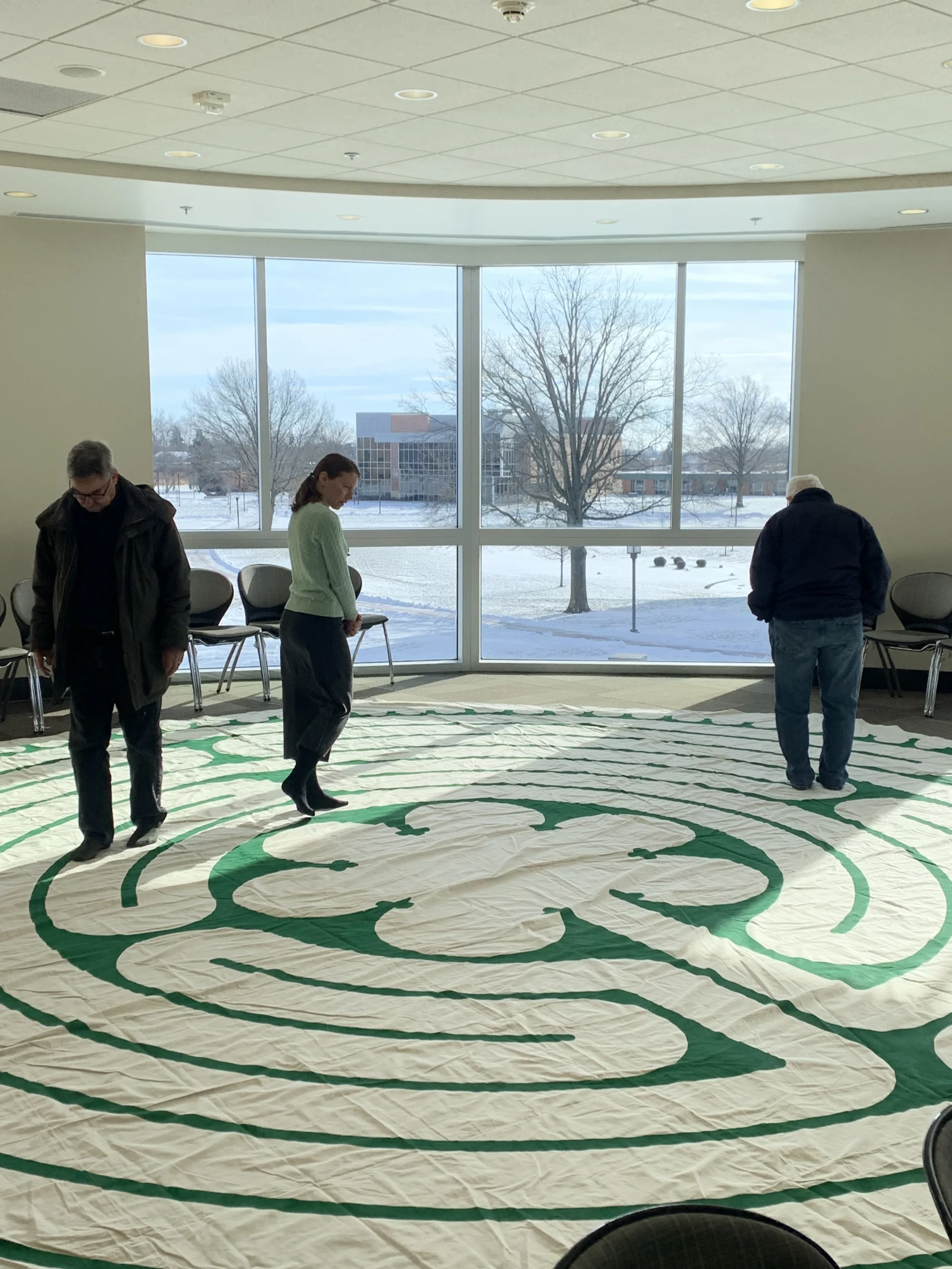 Three people walk on a large, green and white labyrinth mat in a bright room at the Institute for Leadership and Service, with snowy trees visible outside.