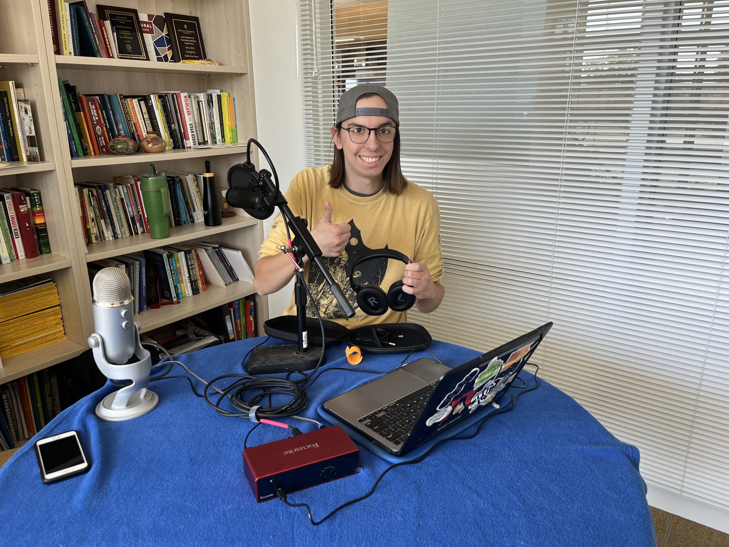 Person smiling at a table with podcast equipment, a laptop, microphone, and headphones in a home office, preparing to record an episode for the Institute for Leadership and Service.