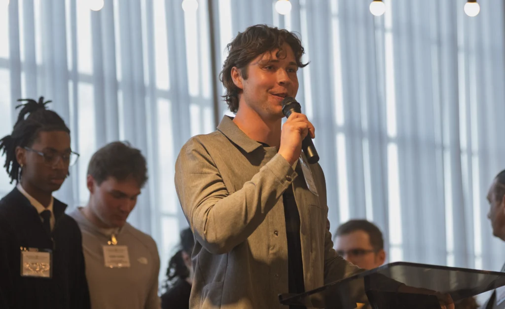 A young Valparaiso University student entrepreneur speaks into a microphone at a podium, with people standing behind him in a bright, well-lit room.