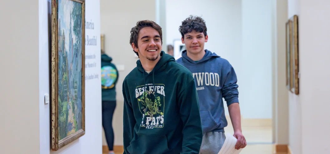 Two teenagers in hoodies smile as they walk through the Brauer Museum of Art, surrounded by framed paintings on the gallery walls.