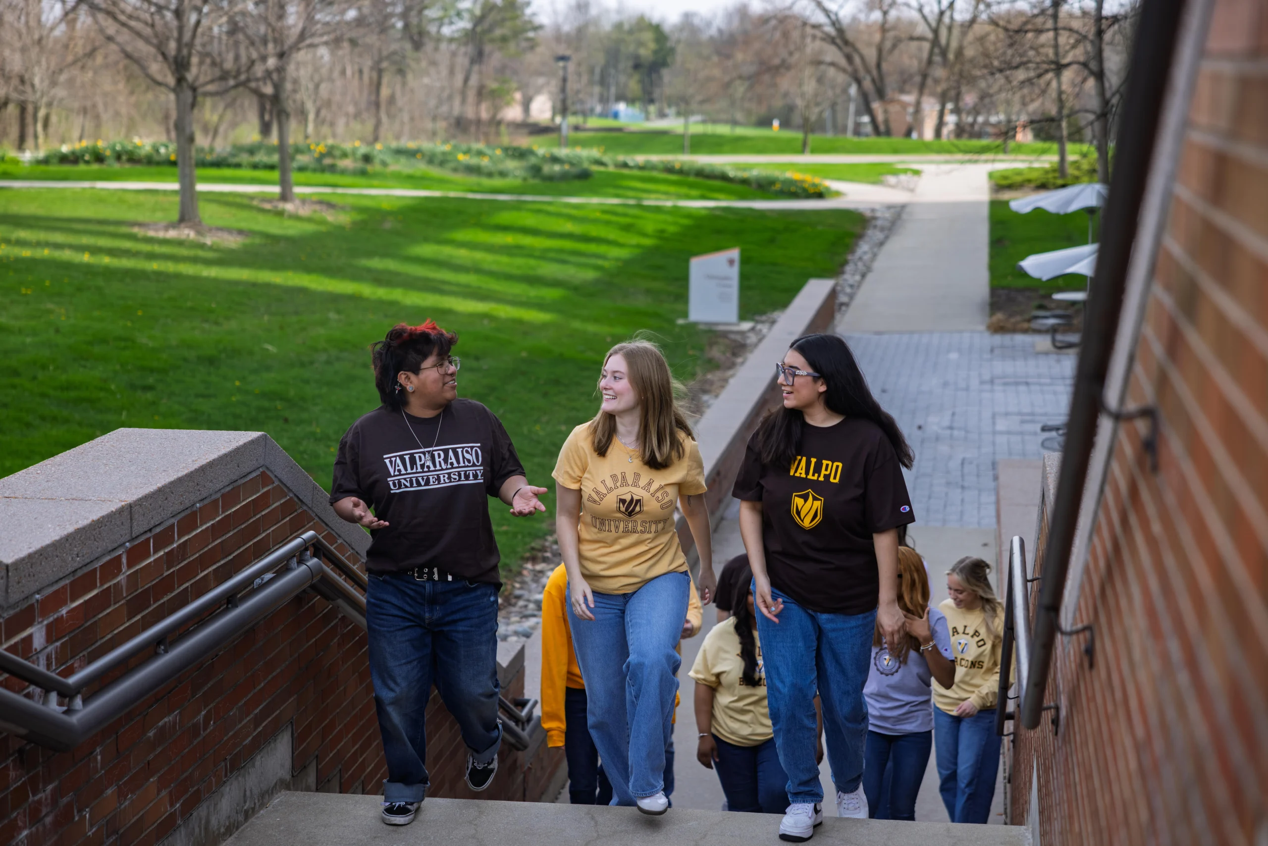 Three transfer students in Valparaiso University shirts walk up outdoor stairs, talking and smiling on a campus pathway.