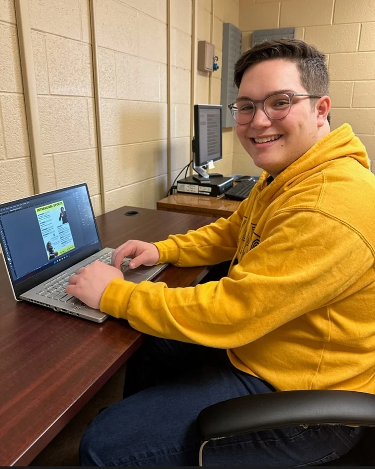 A person in a yellow hoodie smiles while working on a laptop at a desk in an office setting, perhaps researching weather forecasts or following insights from weather profession expert Daniel Murphy.
