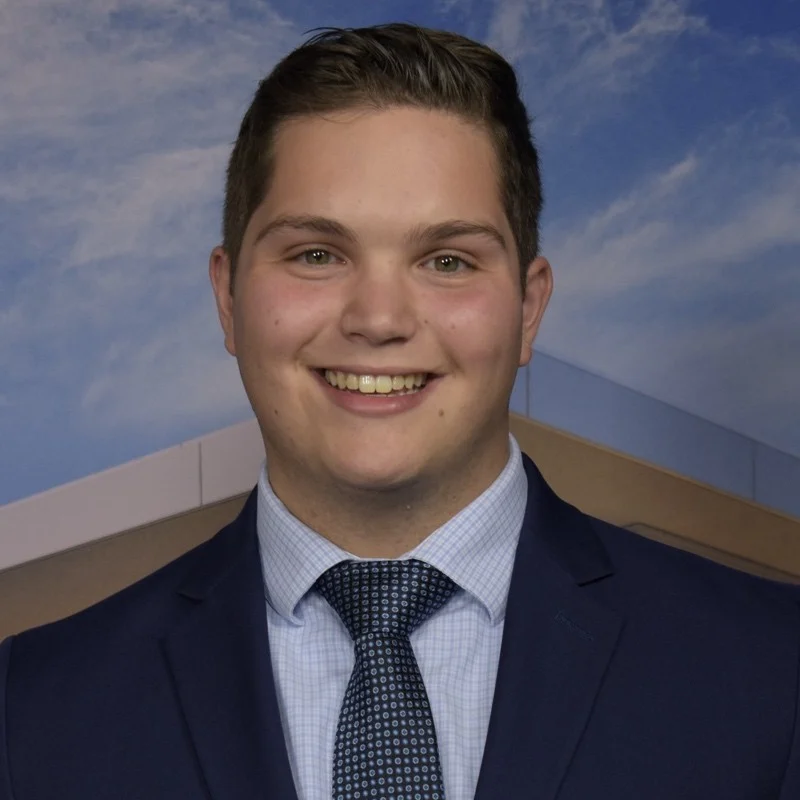 Smiling young man in a suit and tie, possibly Daniel Murphy, poses in front of a blue sky and building background, reflecting his weather profession and expertise in weather forecasts.