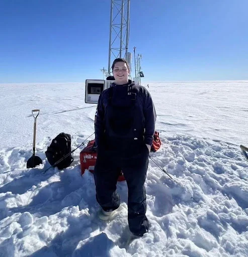 Bundled up in winter gear, Daniel Murphy stands in deep snow near equipment and an antenna, ready to deliver precise weather forecasts under a bright blue sky.