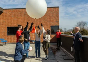 A group of people, including weather profession expert Daniel Murphy, release a large white weather balloon on a sunny rooftop while others watch and take photos.