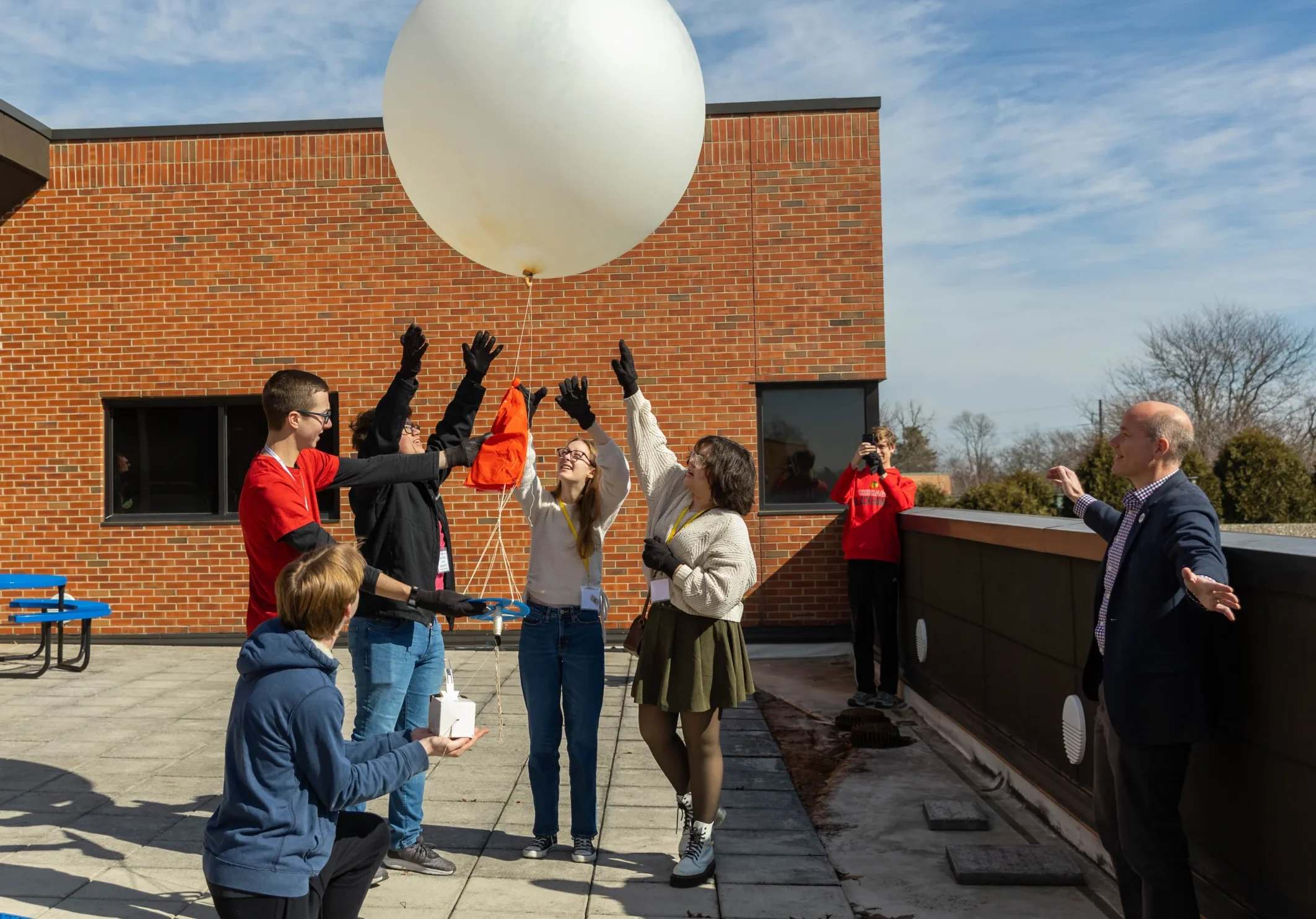 A group of people, including weather profession expert Daniel Murphy, release a large white weather balloon on a sunny rooftop while others watch and take photos.