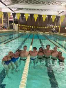 Eight people in swimsuits, including up-and-coming career swimmer Ricky Helmboldt, smile together in an indoor swimming pool under yellow pennant flags.