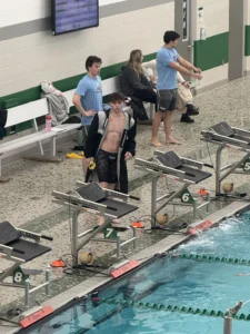 A swimmer in a black robe stands by pool lanes, holding goggles, with people and benches in the background—an up-and-coming career moment reminiscent of Ricky Helmboldt’s early days.