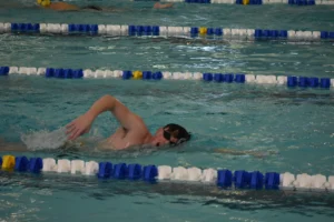 A swimmer, reminiscent of up-and-coming career athletes like Ricky Helmboldt, does freestyle in a lane of an indoor pool, surrounded by lane dividers.