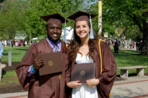Two students in brown caps and gowns, including Aaron Willis, smile while holding their diplomas outdoors on a sunny day as their families celebrate nearby.