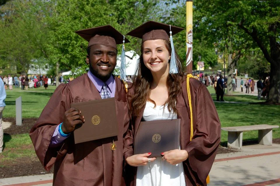 Two students in brown caps and gowns, including Aaron Willis, smile while holding their diplomas outdoors on a sunny day as their families celebrate nearby.