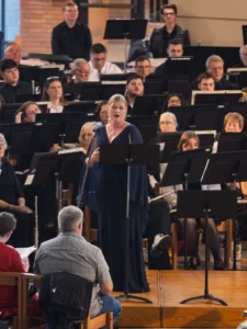 A woman sings with a large orchestra and conductor in a church, celebrating the Sousa Legacy as an audience watches.