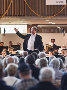 A conductor leads an orchestra performing for a seated audience in an indoor venue, celebrating Sousa’s legacy with a stirring musical tribute.
