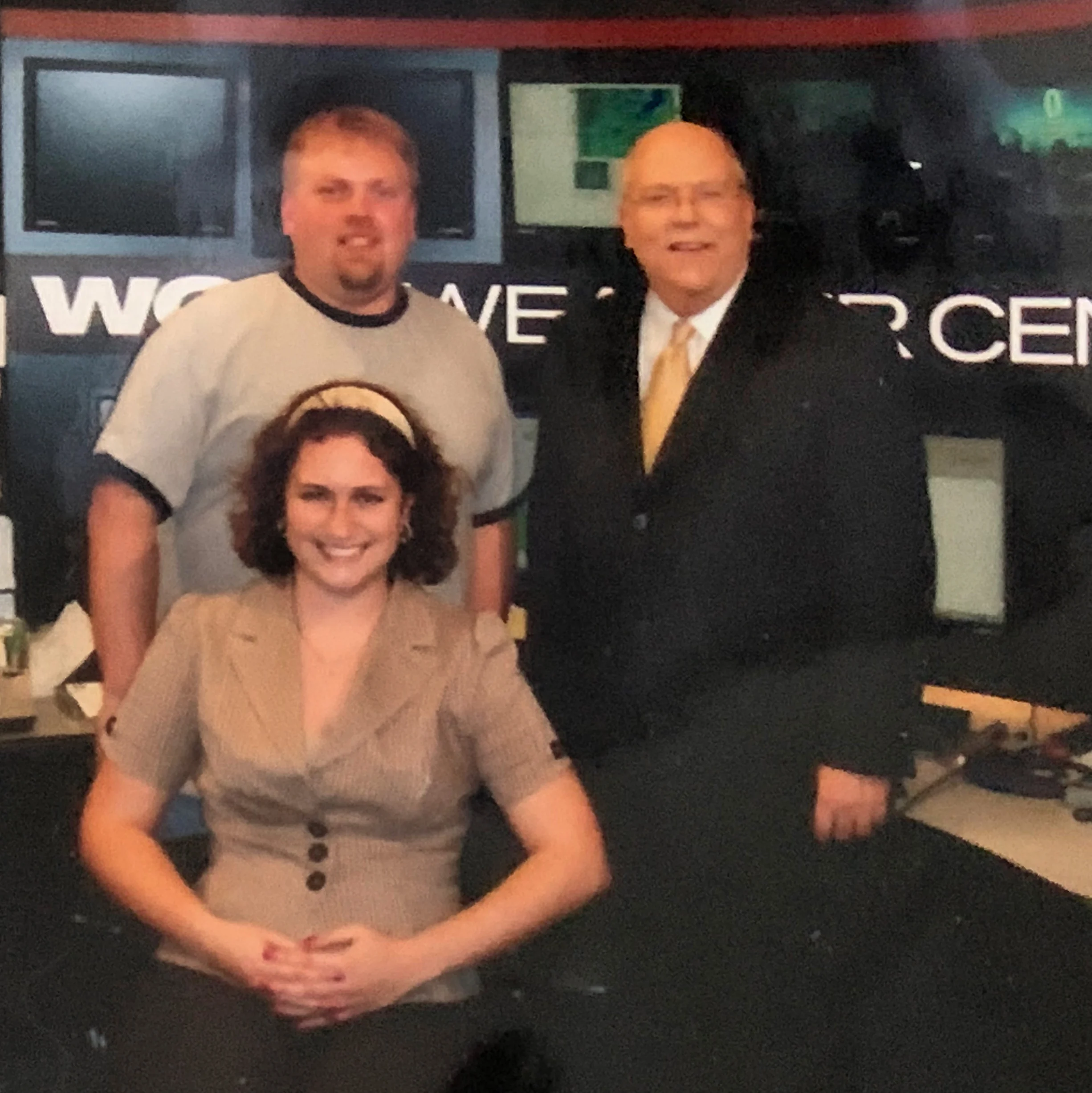 Three people pose and smile in a news studio with monitors and "Weather Center" in the background.