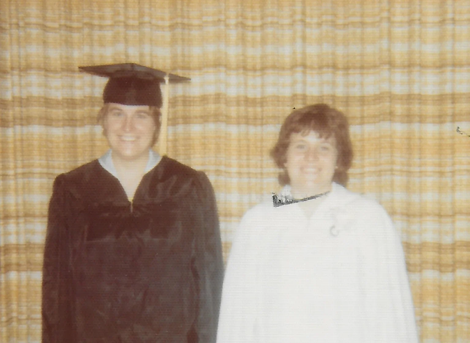 Two people smile, one in a black graduation gown and cap, the other in a white robe, standing indoors at Williams College.