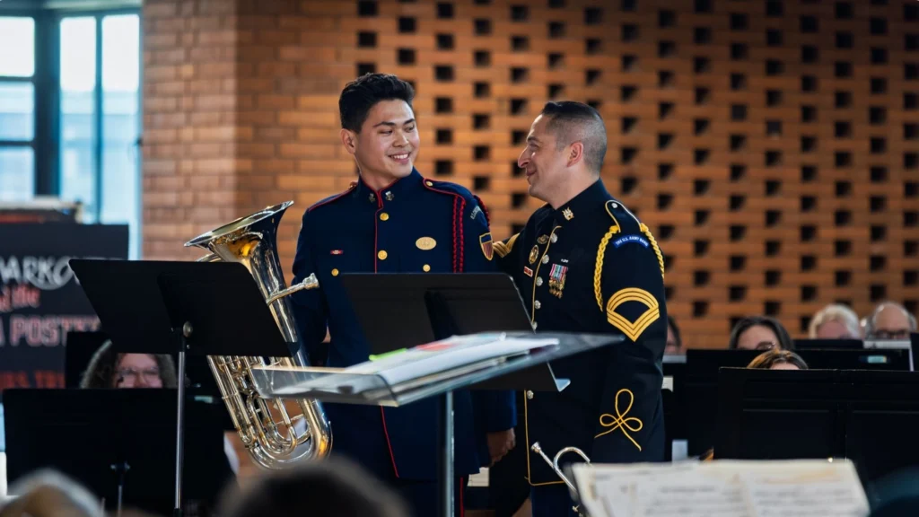 Two uniformed men, one holding a tuba, smile at each other on stage during a band performance, capturing the spirit of the Sousa Legacy.