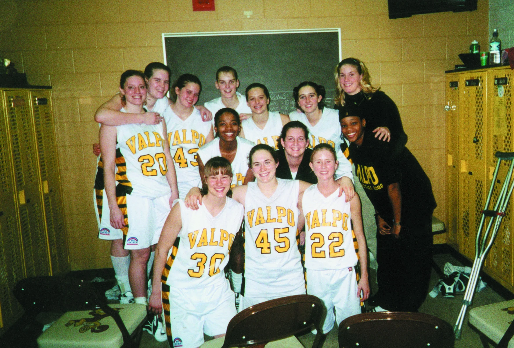 A women’s basketball team in Valpo uniforms poses and smiles together in a locker room, celebrating their win under the guidance of coach Jeanette Gray, a proud Beacon and M.Ed. graduate.