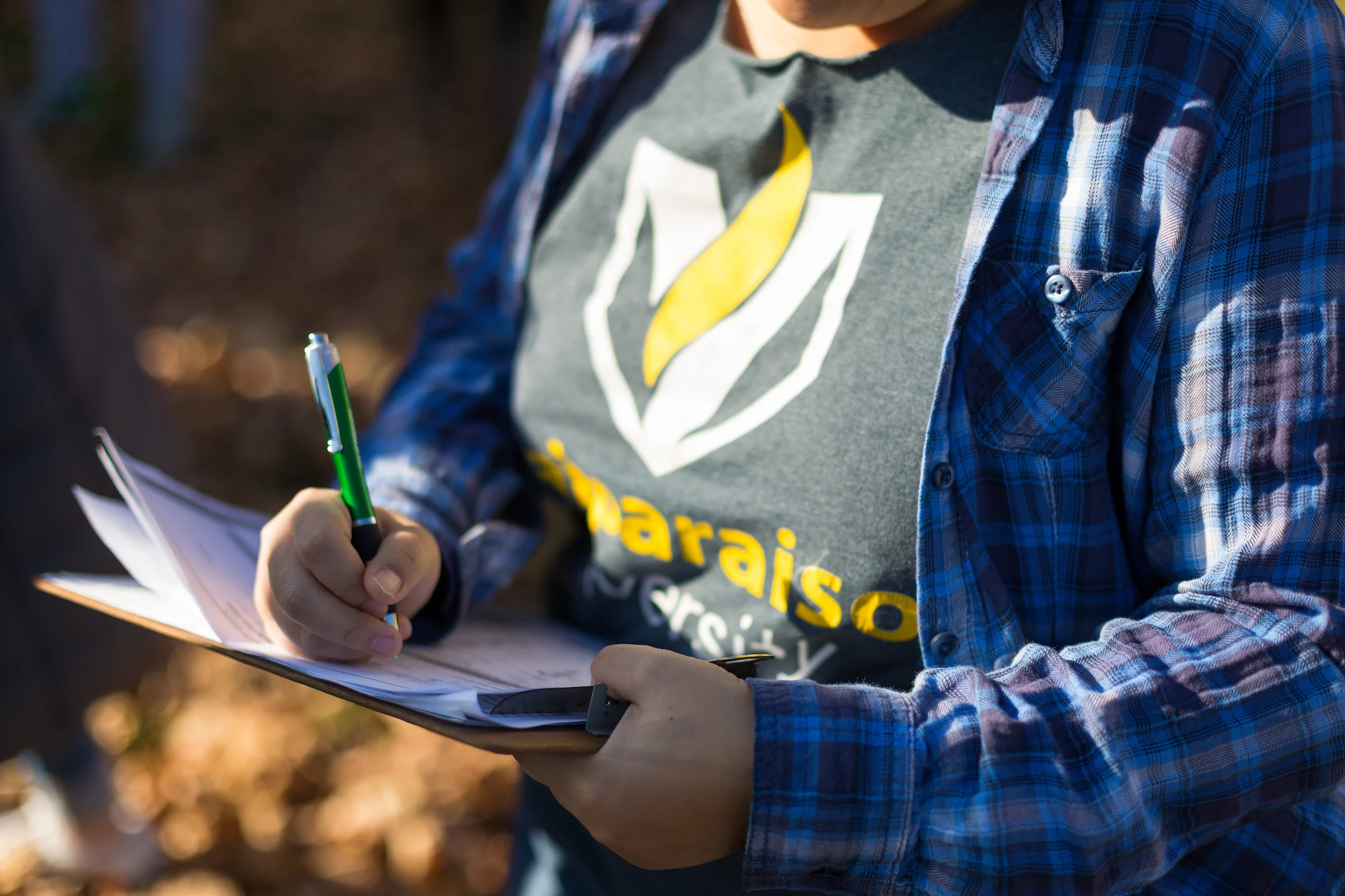 Person in a plaid shirt writing on a clipboard outdoors, holding a green pen and papers.