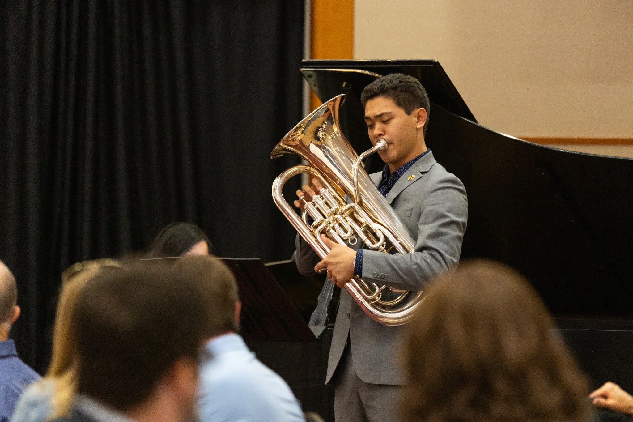 Matthew Yee, the 2024 Tau Beta Pi Laureate, plays a euphonium on stage in front of an audience and a black piano, dressed smartly in a suit.