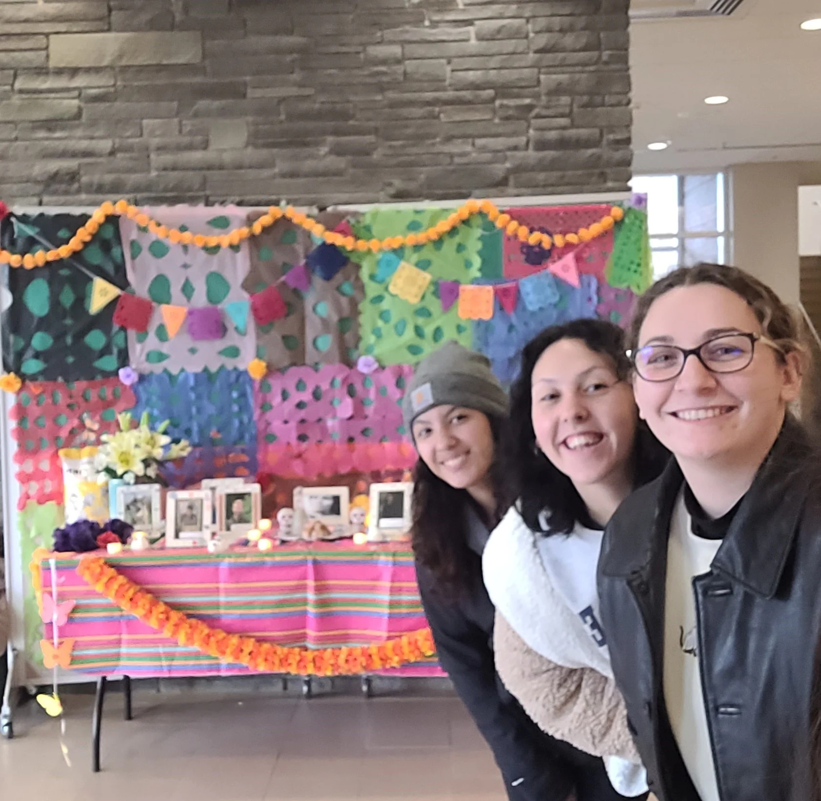 Three smiling people pose in front of a colorful, decorated table with framed photos and festive streamers, celebrating the vibrant spirit of Multicultural Programs.