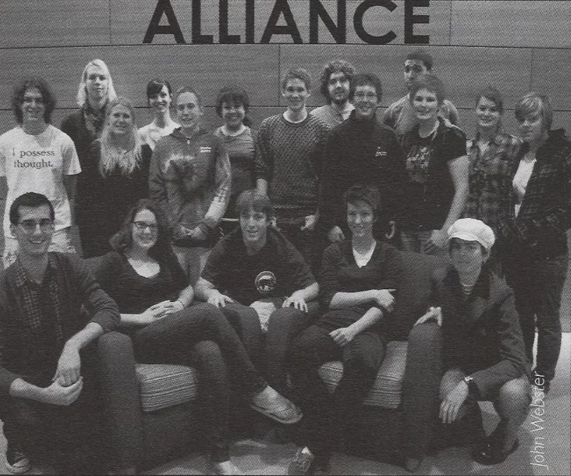 A group of 19 people posing together indoors in front of a wall with the word "ALLIANCE" above them, including Gideon Litherland at the center, creating a beacon of unity and collaboration.