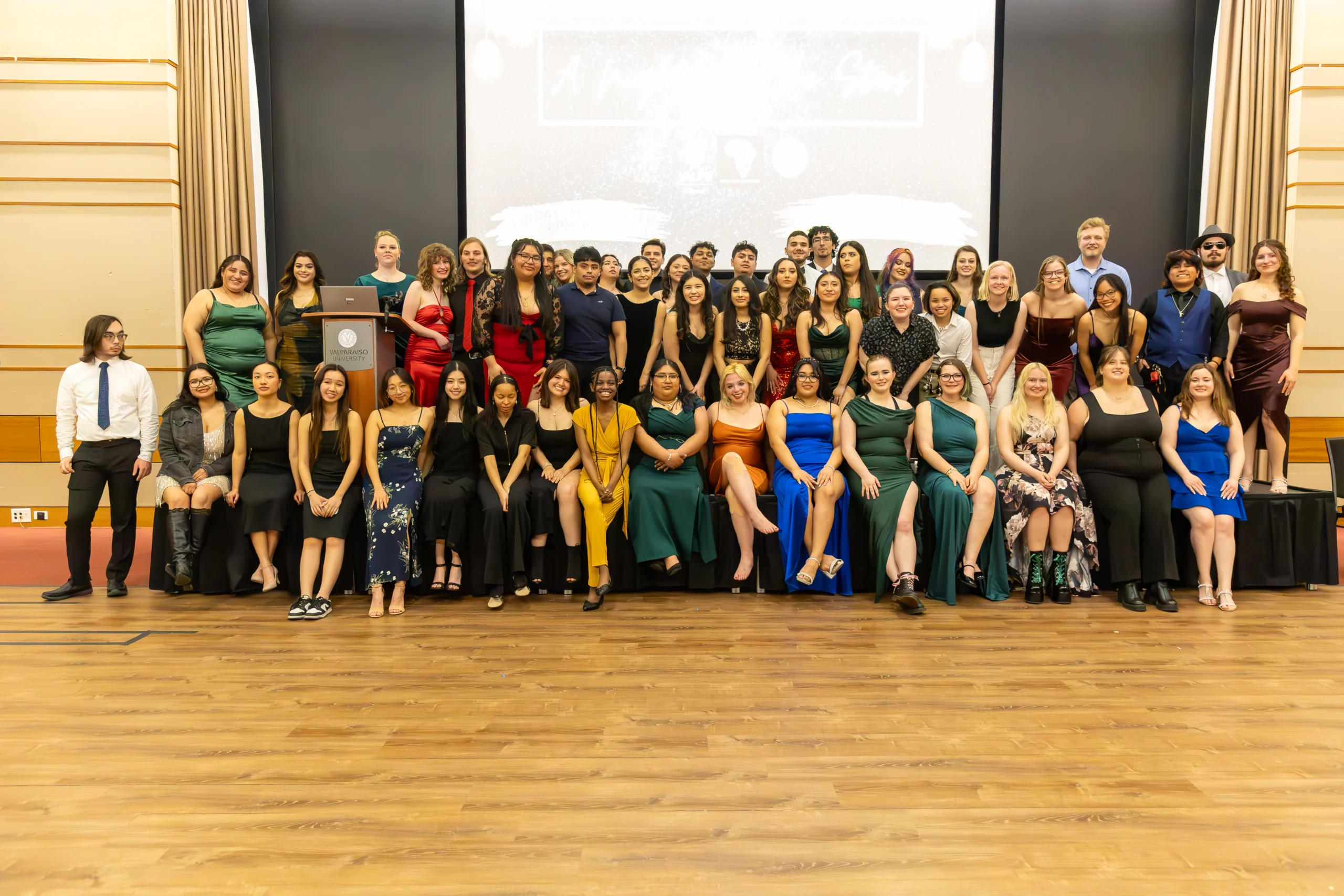 A large group of people in formal attire pose together on a stage in a ballroom, representing various multicultural organizations.