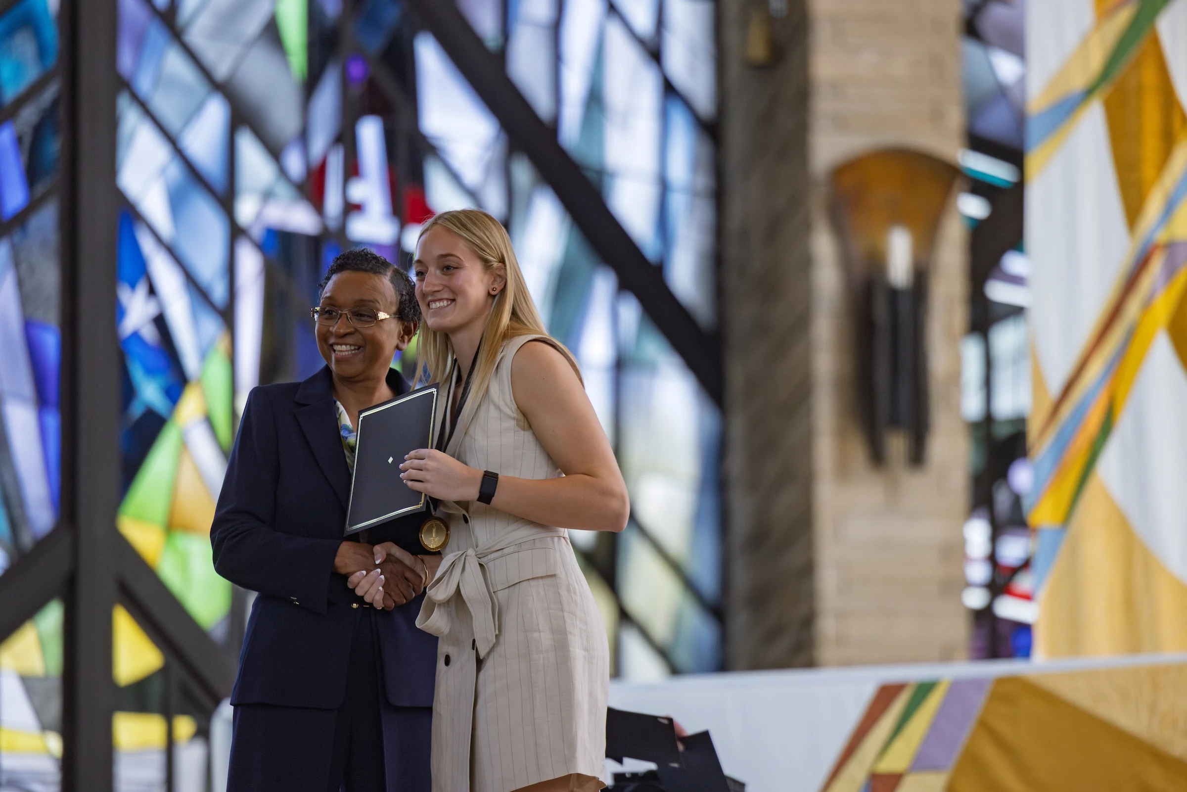 Two women smiling and posing with an award in front of colorful stained glass windows, celebrating a professional opportunity.