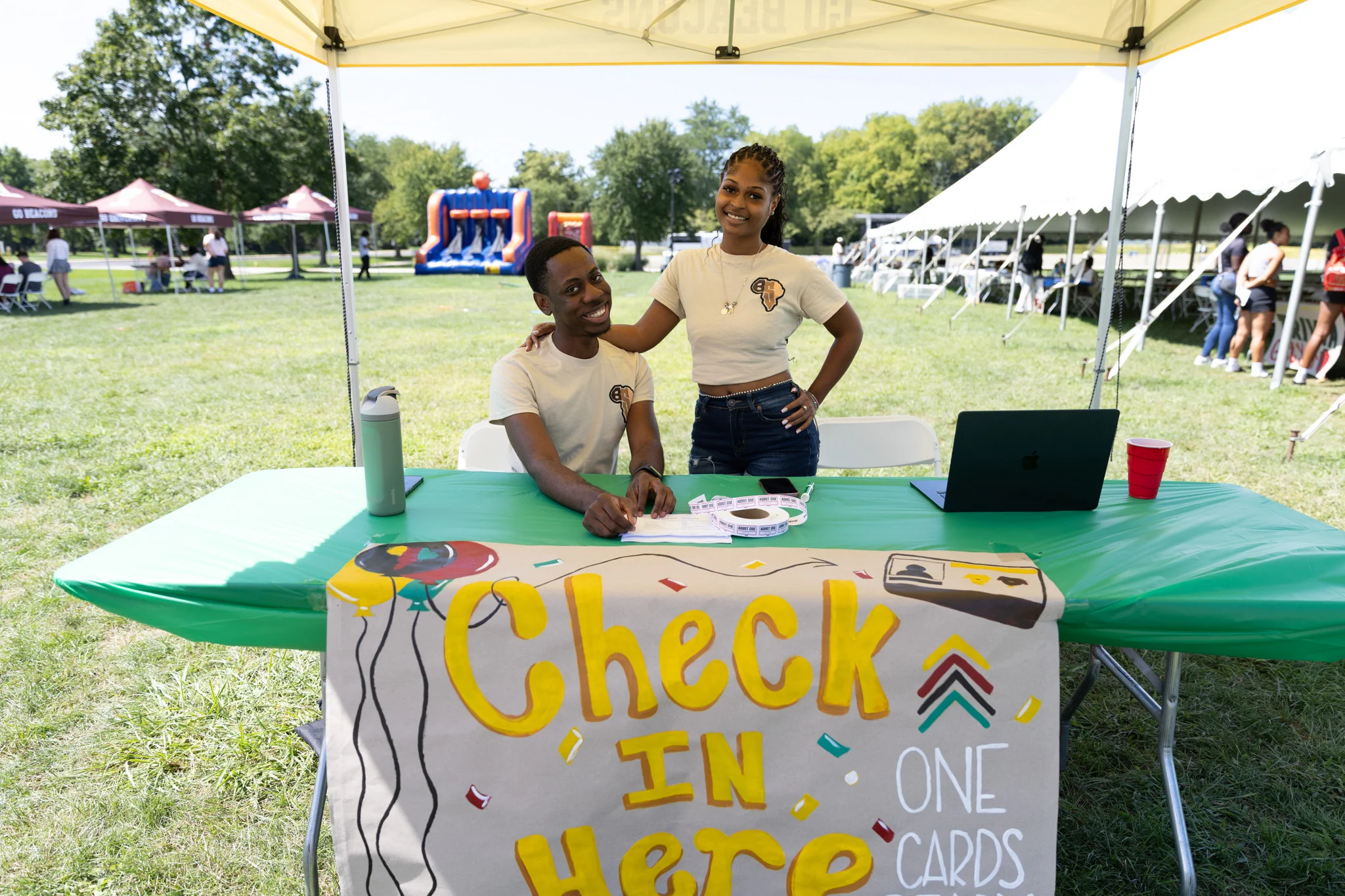 Two people at an outdoor check-in table with a sign for Multicultural Organizations, a laptop, and tents in the background.