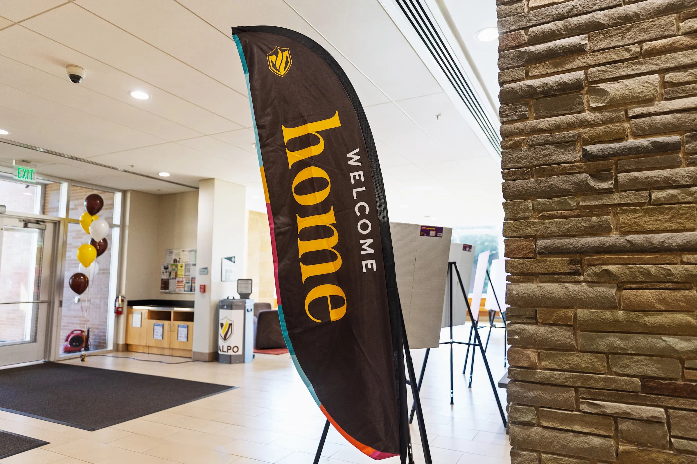 Tall feather flag with bold typography in a bright lobby reads “Welcome Home”; balloons and a stone wall are visible nearby.