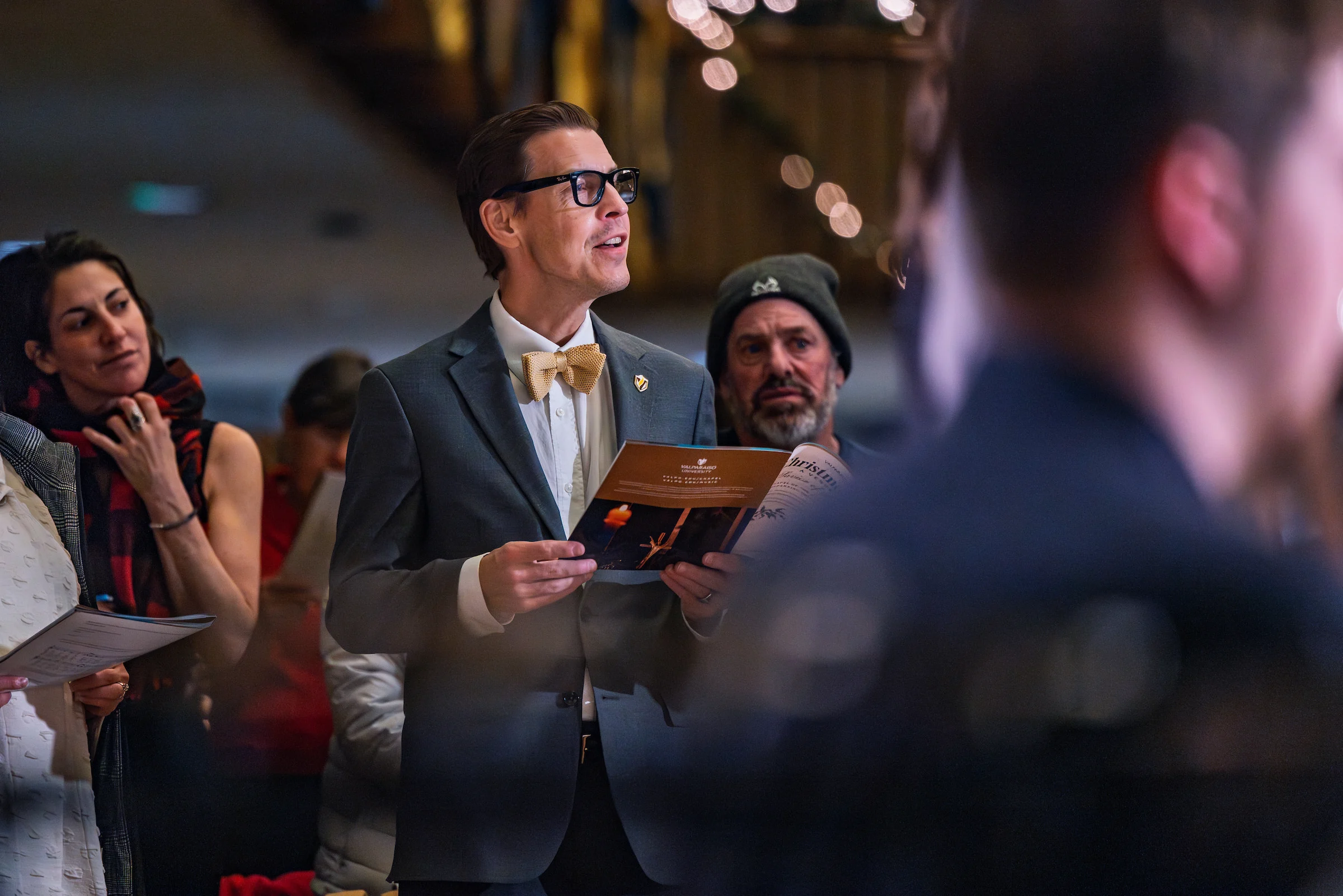 Man in a suit and bow tie holding a program, standing among others at an indoor event organized by the President's Office.
