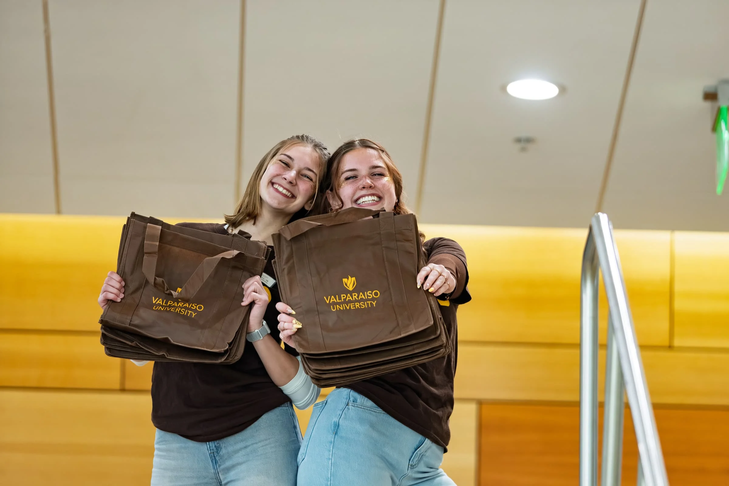 Two smiling students hold Valparaiso University tote bags while standing on a staircase indoors, reflecting the spirit found in our Brand Style Guides.