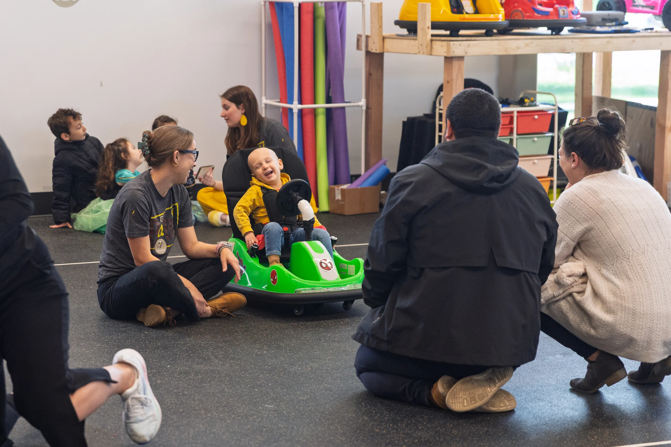 A smiling child enjoys newfound mobility as they ride a green GoBabyGo toy car indoors, surrounded by laughing adults and other children.