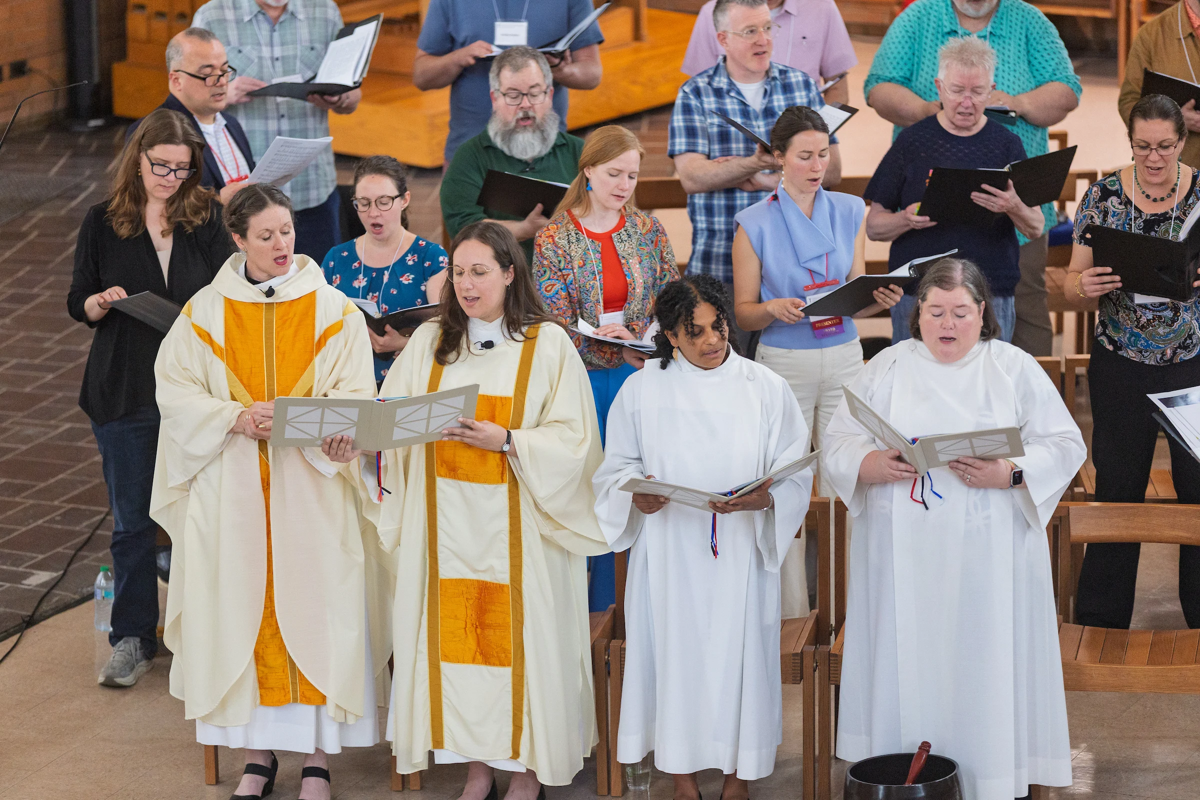 A choir sings in a church, with three women in white robes and others standing behind holding music sheets during the Institute of Liturgical Studies.