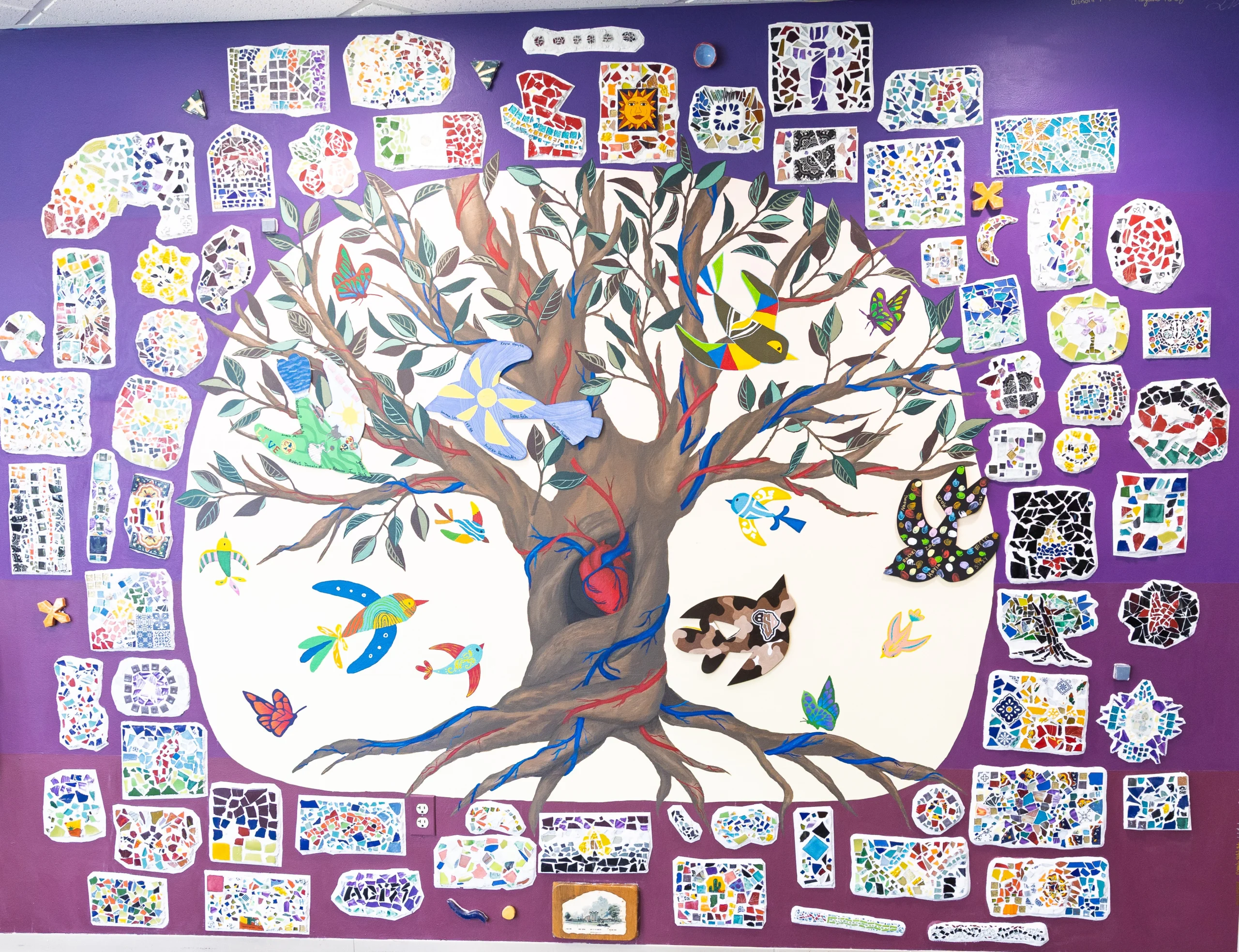 Colorful mural of a tree with birds, surrounded by mosaic art pieces on a bright purple wall, celebrating Multicultural Programs and diverse communities.