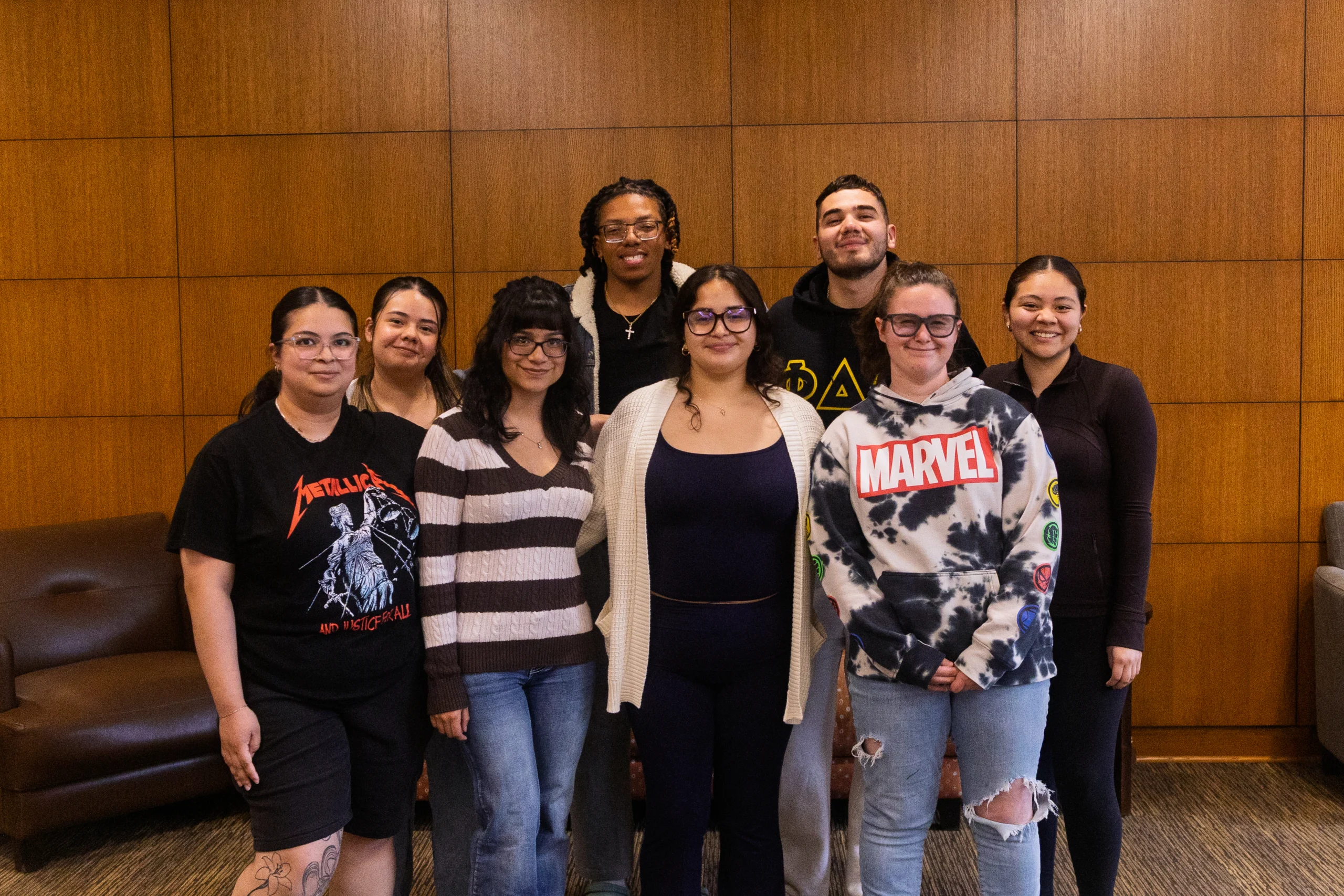 A group of eight young adults stands together indoors, smiling in front of a wooden wall, proudly representing the Cornerstone Program.