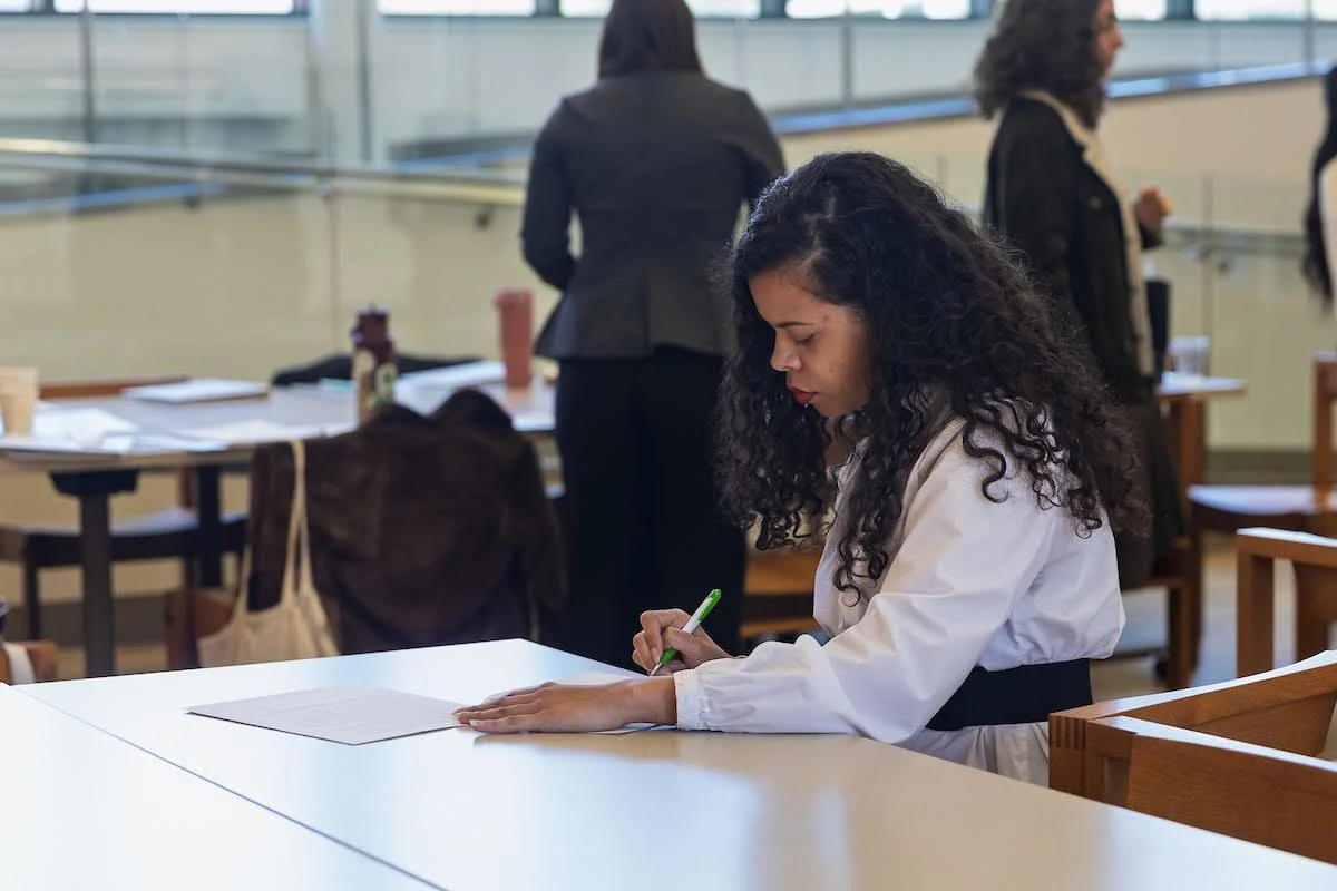 Woman with curly hair writing on paper at a table in a bright room at Valpo, making a community impact while others engage in the background.