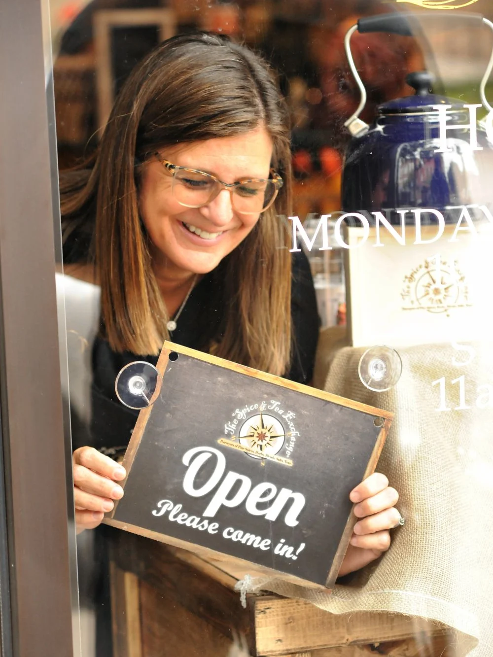 Smiling woman places an "Open, Please come in!" sign in the window of a cozy St. Aubin shop.
