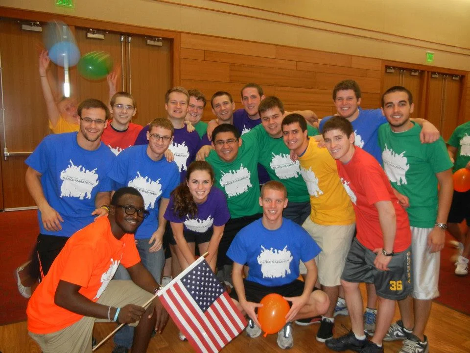 A group of young adults in colorful shirts pose indoors, smiling, with one person holding an American flag like a beacon of unity—among them stands Aaron Willis.