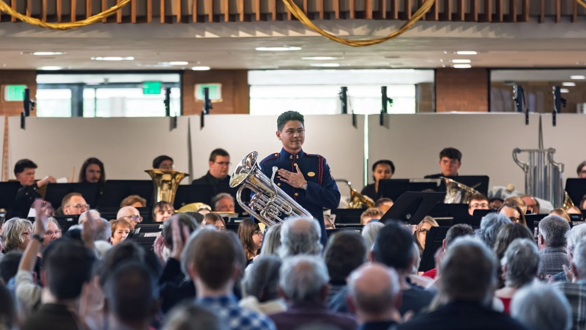 A young musician holding a euphonium stands and smiles as an audience applauds, celebrating a dream performance during a band concert.